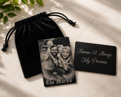 Two black aluminum wallet cards engraved with a family photograph and custom message beside a black velvet pouch on a cream stone surface.