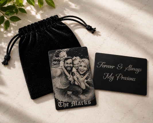 Two black aluminum wallet cards engraved with a family photograph and custom message beside a black velvet pouch on a cream stone surface.